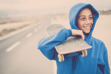 Lächelnder Junge mit Skateboard auf der Schulter auf leerer Landstraße - SIPF00941