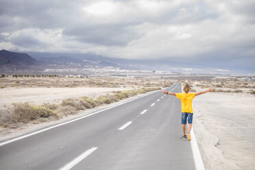 Spanien, Teneriffa, Rückenansicht eines Jungen mit ausgestreckten Armen, der auf einer leeren Landstraße läuft - SIPF00942