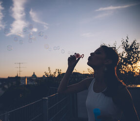 Young woman blowing soap bubbles on roof terrace in the evening twilight - AIF00408