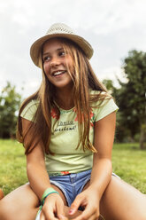 Porträt eines lächelnden Mädchens mit Sommerhut auf einer Wiese sitzend - MGOF02553