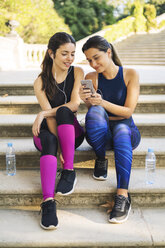Zwei sportliche junge Frauen sitzen auf einer Treppe und schauen auf ihr Handy - EBSF01793