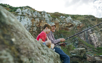 Lächelnder Großvater und Enkel fischen zusammen auf einem Felsen sitzend - DAPF00426