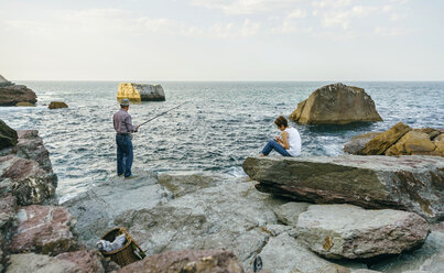 Senior man fishing at the sea with wife sitting on rock - DAPF00434