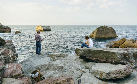 Älterer Mann beim Fischen am Meer mit Frau auf einem Felsen sitzend - DAPF00434