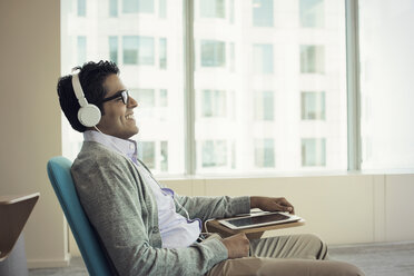 Businessman sitting in chair, wearing headphones, using digital tablet - WESTF21765