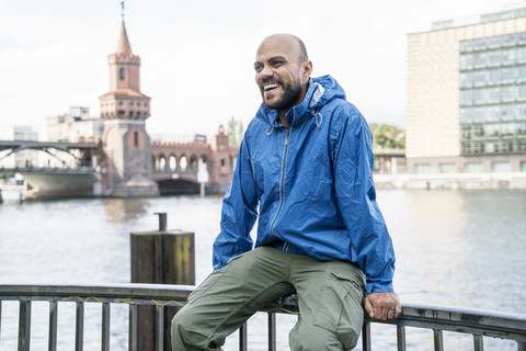 Deutschland, Berlin, lachender Mann in blauer Regenjacke sitzt auf dem Geländer vor der Oberbaumbrücke, lizenzfreies Stockfoto