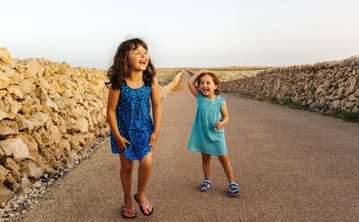 Zwei lachende kleine Schwestern stehen auf leerer Straße bei Sonnenuntergang - MGOF02567
