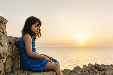 Trauriges kleines Mädchen, das bei Sonnenuntergang vor dem Meer sitzt - MGOF02568