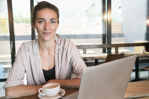 Portrait of smiling young woman with cup of coffee and laptop in a coffee shop - TAMF00731