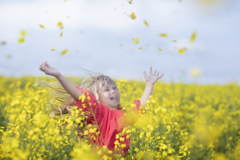 Fröhliches kleines Mädchen steht im Rapsfeld und wirft Blüten in die Luft, lizenzfreies Stockfoto