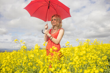 Blond young woman wearing red dress standing in rape field holding red umbrella - ZEF11137