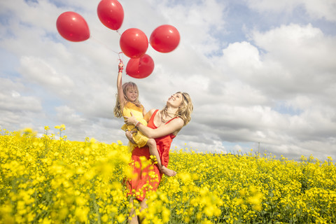 Mutter und kleine Tochter haben Spaß mit roten Luftballons im Rapsfeld, lizenzfreies Stockfoto