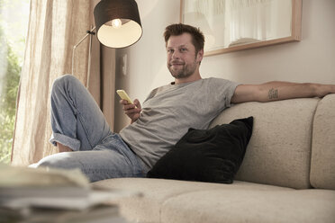 Entspannter Mann zu Hause auf der Couch sitzend mit Mobiltelefon - SUF00088