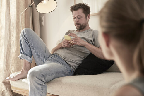 Mann zu Hause auf der Couch sitzend und auf sein Handy schauend mit Frauen im Vordergrund - SUF00089