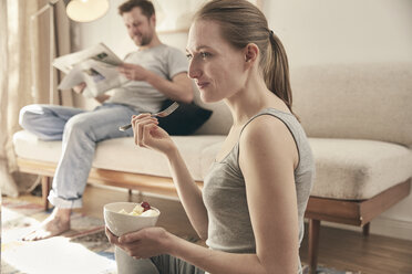 Woman eating fruits in living room with man in background - SUF00091