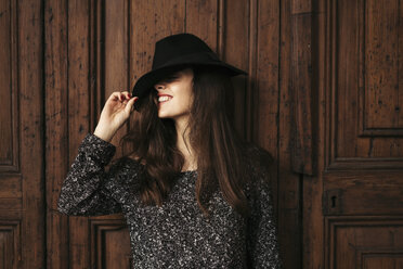 Lächelnde junge Frau steht vor einer Holztür und verdeckt die Augen mit ihrem Hut - EBSF01846