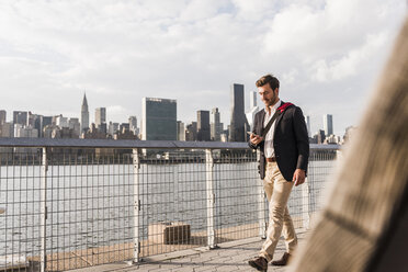 USA, New York City, Geschäftsmann geht am East River entlang und schaut auf sein Handy - UUF08846