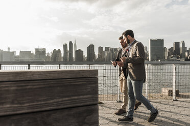 USA, New York City, zwei Geschäftsleute gehen am East River entlang und schauen auf ihr Handy - UUF08854