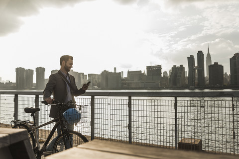 USA, New York City, businessman with bicycle walking along East River looking at cell phone stock photo