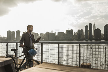 USA, New York City, Geschäftsmann mit Fahrrad geht am East River entlang und schaut auf sein Handy - UUF08872