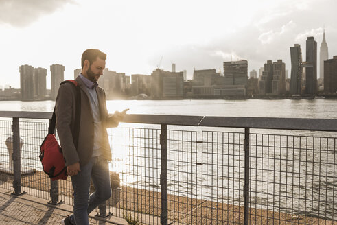 USA, New York City, Geschäftsmann geht am East River entlang und schaut auf sein Handy - UUF08890