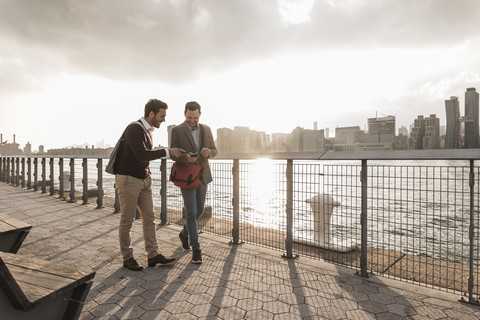 USA, New York City, zwei junge Männer gehen am East River entlang und schauen auf ihr Handy, lizenzfreies Stockfoto