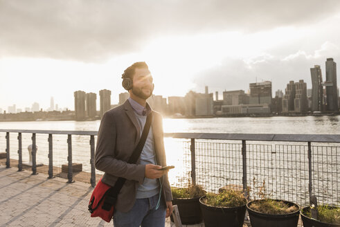 USA, New York City, junger Mann mit Kopfhörern und Mobiltelefon am East River - UUF08909