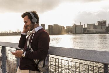 USA, New York City, smiling young man with headphones and cell phone at East River - UUF08913