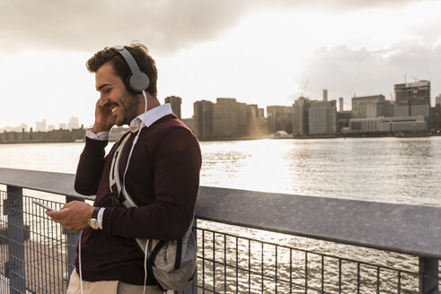 USA, New York City, lächelnder junger Mann mit Kopfhörern und Mobiltelefon am East River - UUF08913