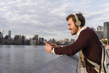 USA, New York City, lächelnder junger Mann mit Kopfhörern und Mobiltelefon am East River - UUF08914