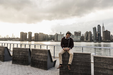 USA, New York City, junger Mann mit Kopfhörern und Mobiltelefon am East River sitzend - UUF08917