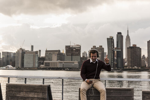 USA, New York City, junger Mann mit Kopfhörern und Mobiltelefon am East River sitzend - UUF08918