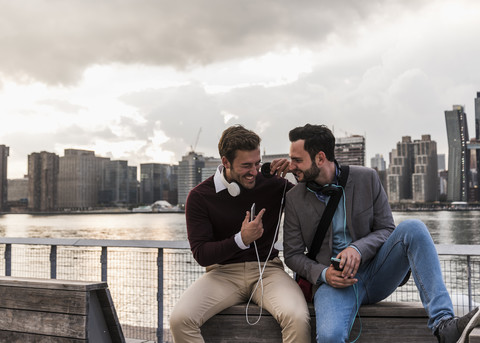 USA, New York City, two happy young men with headphones and cell phone sitting at East River stock photo