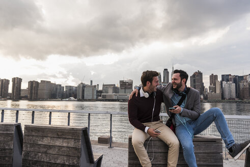 USA, New York City, zwei glückliche junge Männer mit Kopfhörern und Handy sitzen am East River - UUF08922