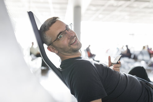 Mann sitzt im Abflugbereich eines Flughafens und hält ein Smartphone in der Hand - SHKF00693