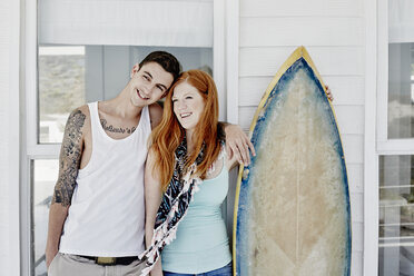 Glückliches junges Paar mit Surfbrett vor einem Strandhaus stehend - RORF00371
