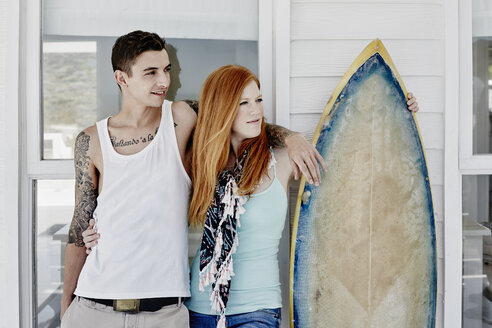 Glückliches junges Paar mit Surfbrett vor einem Strandhaus stehend - RORF00372