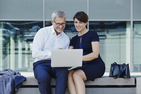 Geschäftsmann und Geschäftsfrau sitzen auf einer Bank und teilen sich einen Laptop, lizenzfreies Stockfoto