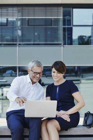 Geschäftsmann und Geschäftsfrau sitzen auf einer Bank und teilen sich einen Laptop, lizenzfreies Stockfoto