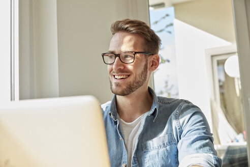 Portrait of smiling man with laptop at home looking through window - FMKF03134