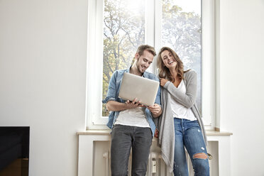 Glückliches Paar vor einem Fenster stehend mit Laptop - FMKF03139
