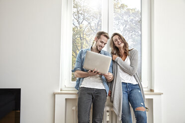 Happy couple standing in front of window with laptop - FMKF03139