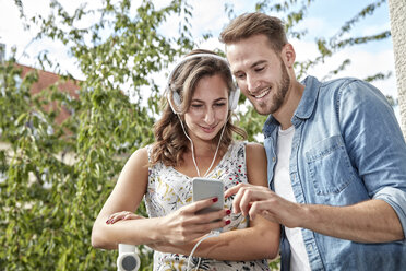 Lächelndes Paar auf dem Balkon mit Kopfhörern und Smartphone - FMKF03147