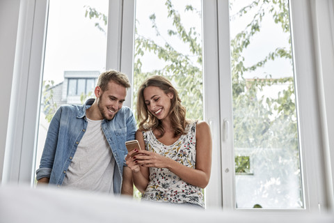 Lächelndes Paar vor einem Fenster mit Blick auf ein Smartphone, lizenzfreies Stockfoto