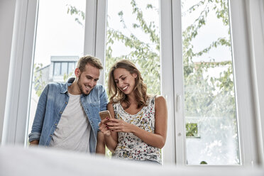 Lächelndes Paar vor einem Fenster mit Blick auf ein Smartphone - FMKF03151