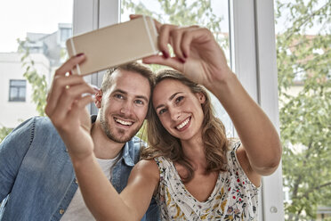 Porträt eines lächelnden Paares vor einem Fenster, das ein Selfie mit einem Smartphone macht - FMKF03152