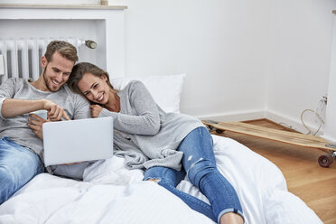Smiling couple lying on bedding on the floor looking at laptop - FMKF03153