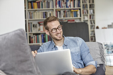 Porträt eines lächelnden Mannes auf der Couch mit Laptop im Wohnzimmer - FMKF03154