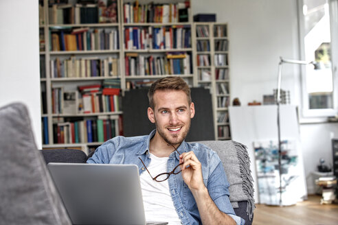 Portrait of smiling man sitting on couch with laptop in the living room - FMKF03155