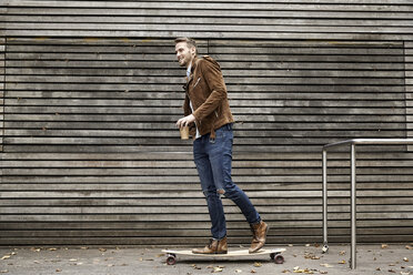 Lächelnder Mann mit Kaffee zum Mitnehmen auf dem Skateboard vor einer Holzwand - FMKF03160